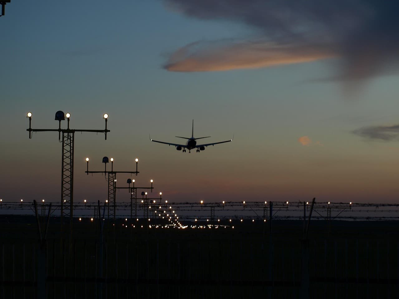 An airliner on final approach at dusk, low over the runway approach lights